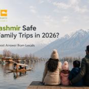 A family sits beside Dal Lake in Kashmir with snow-covered mountains and a traditional shikara boat in the background, alongside the headline Is Kashmir Safe for Family Trips in 2026? The Honest Answer from Locals